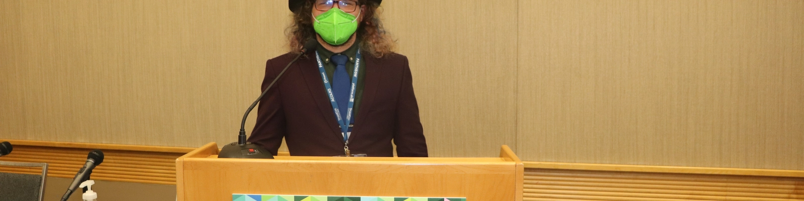 Nick speaking at DrupalCon North America Portland 2022 -  behind a lecturn with DrupalCon signage, wearing attendee badge over purple suit with a green mask and a black hat.