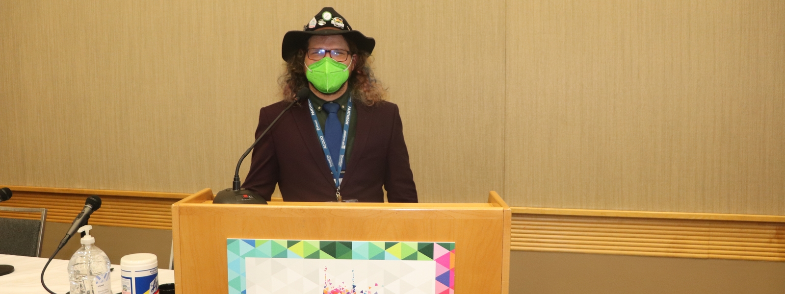 Nick speaking at DrupalCon North America Portland 2022 -  behind a lecturn with DrupalCon signage, wearing attendee badge over purple suit with a green mask and a black hat.