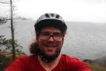 Nick wearing a red shirt and a bike helmet with cloud and wind swept ocean behind him